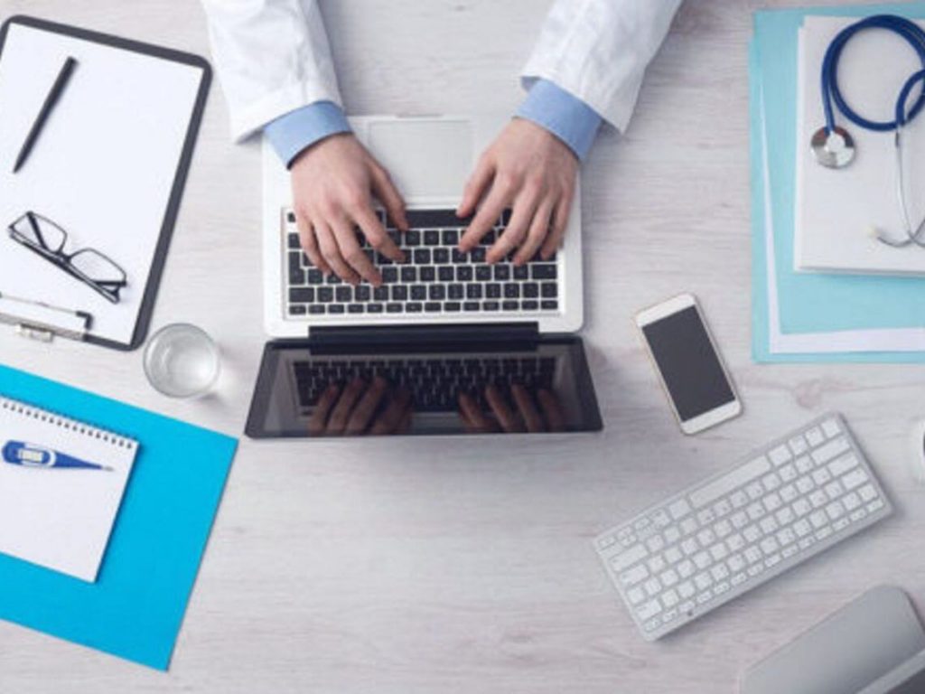 A doctor is working on his laptop at his desk with different technologies on the table