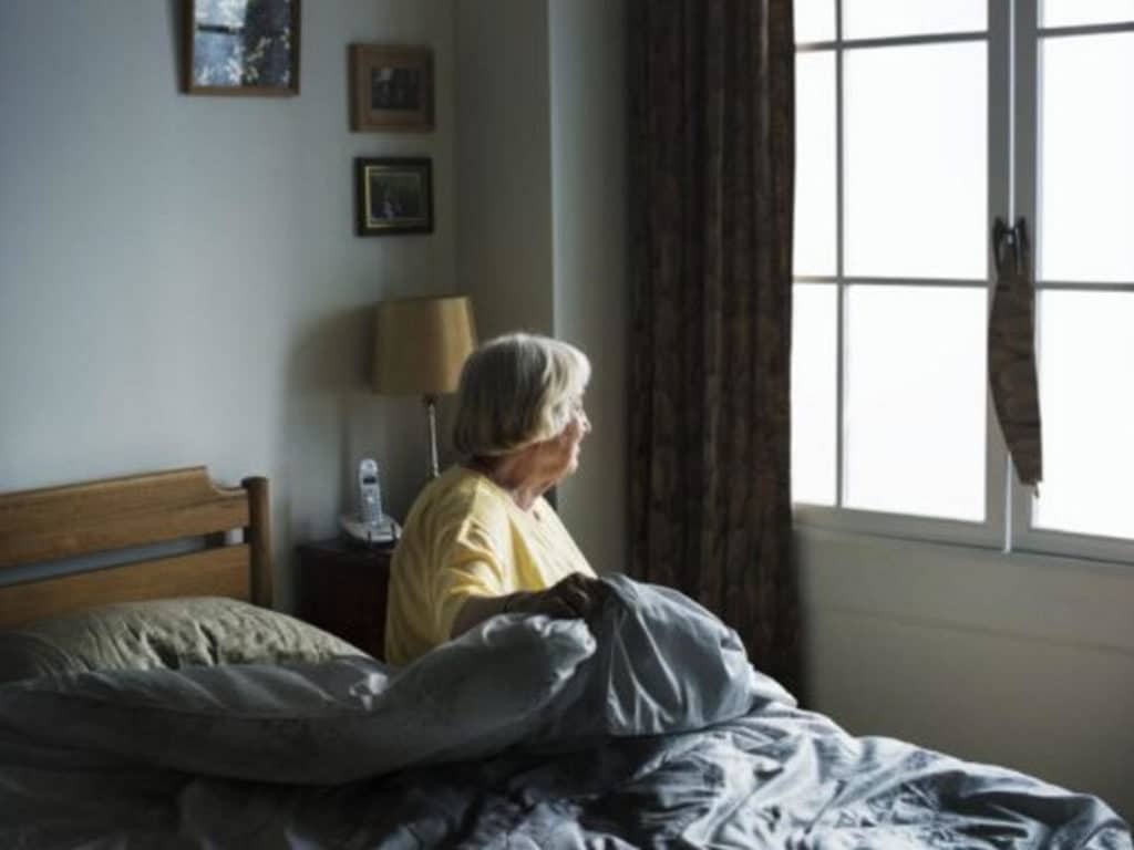 An elderly woman sits in her bed looking out of her window