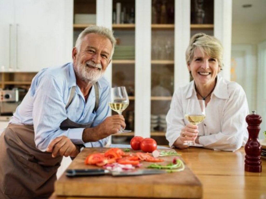 A man and a woman are drinking wine over a kitchen counter full of food